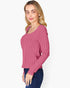 A woman modeling a pink ribbed knit long-sleeve top with a scoop neckline, paired with dark blue jeans, against a plain light gray background.