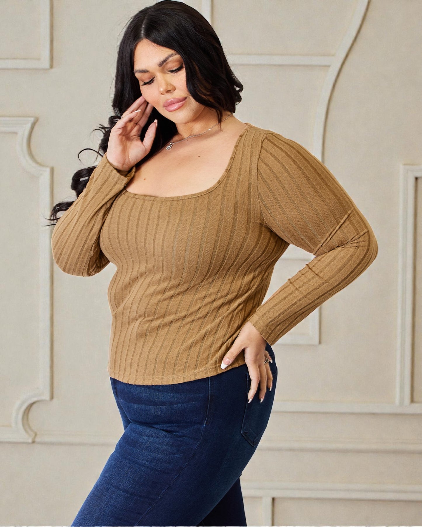 Woman wearing a brown ribbed sweater and blue jeans against a beige wall.