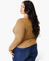 A woman with long, wavy dark hair is shown from a side-back angle wearing a tan ribbed long-sleeve top and dark blue jeans against a plain white studio background.
