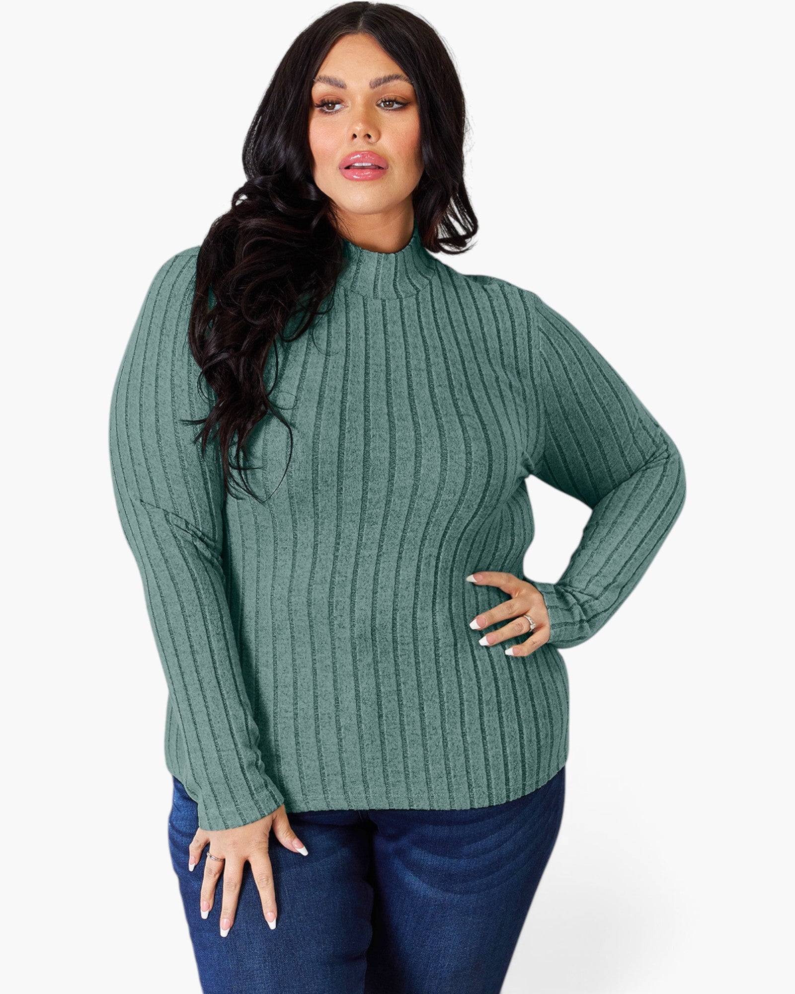 A woman modeling a ribbed knit mock neck sweater in sage green, paired with dark blue jeans, against a plain white studio background.