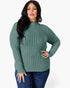 A woman modeling a ribbed knit mock neck sweater in sage green, paired with dark blue jeans, against a plain white studio background.