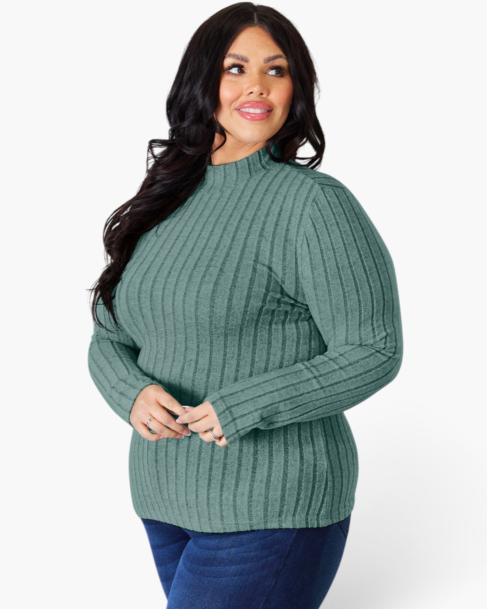 A woman modeling a ribbed knit mock neck sweater in sage green, paired with dark blue jeans, against a plain white studio background.