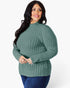 A woman modeling a ribbed knit mock neck sweater in sage green, paired with dark blue jeans, against a plain white studio background.