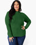 A woman modeling a green ribbed mock neck sweater with long sleeves, paired with blue jeans, against a plain light gray background.
