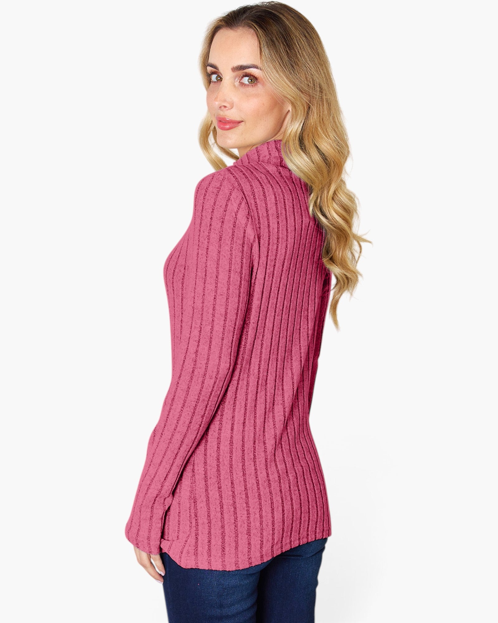 A woman with blonde wavy hair is shown from a side-back angle wearing a pink ribbed mock neck sweater and dark blue jeans against a plain white background.