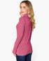 A woman with blonde wavy hair is shown from a side-back angle wearing a pink ribbed mock neck sweater and dark blue jeans against a plain white background.
