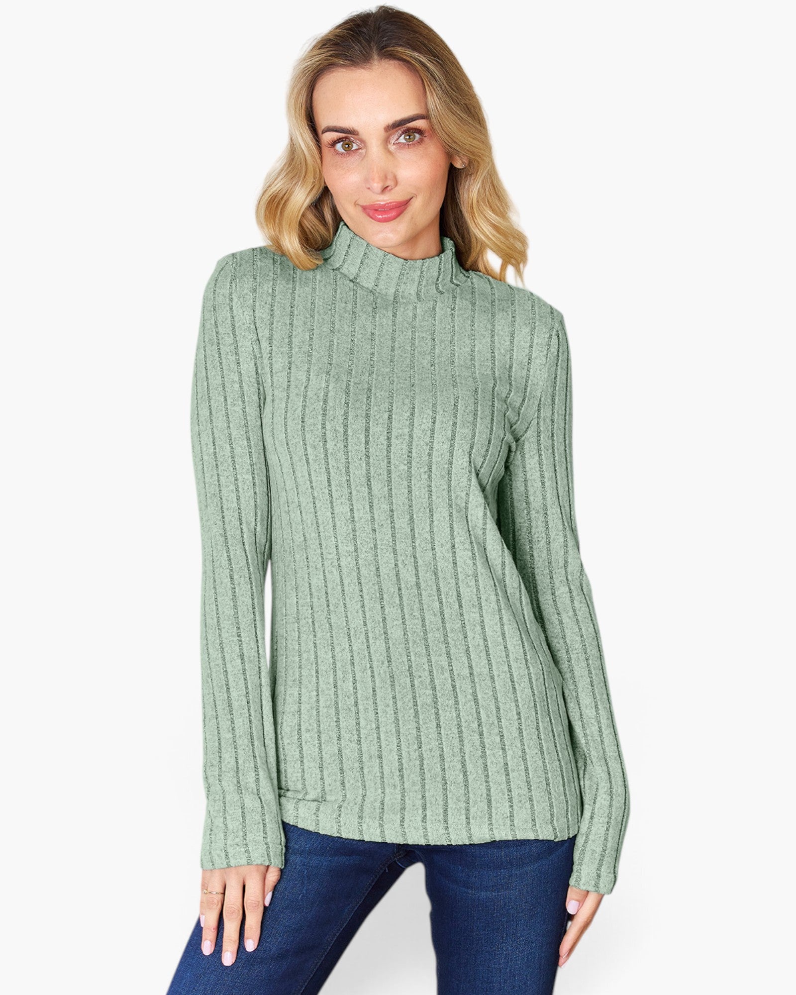 A woman wearing a sage green ribbed mock neck sweater and blue jeans, standing against a plain white studio background.