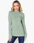 A woman wearing a sage green ribbed mock neck sweater and blue jeans, standing against a plain white studio background.