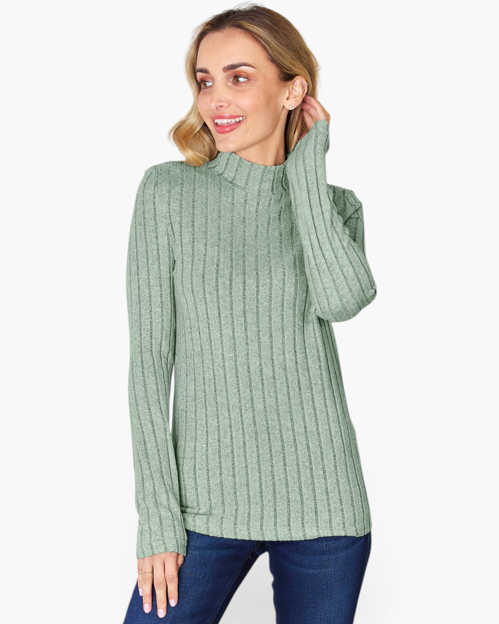 A woman wearing a sage green ribbed mock neck sweater and dark blue jeans, posing against a light gray studio background.