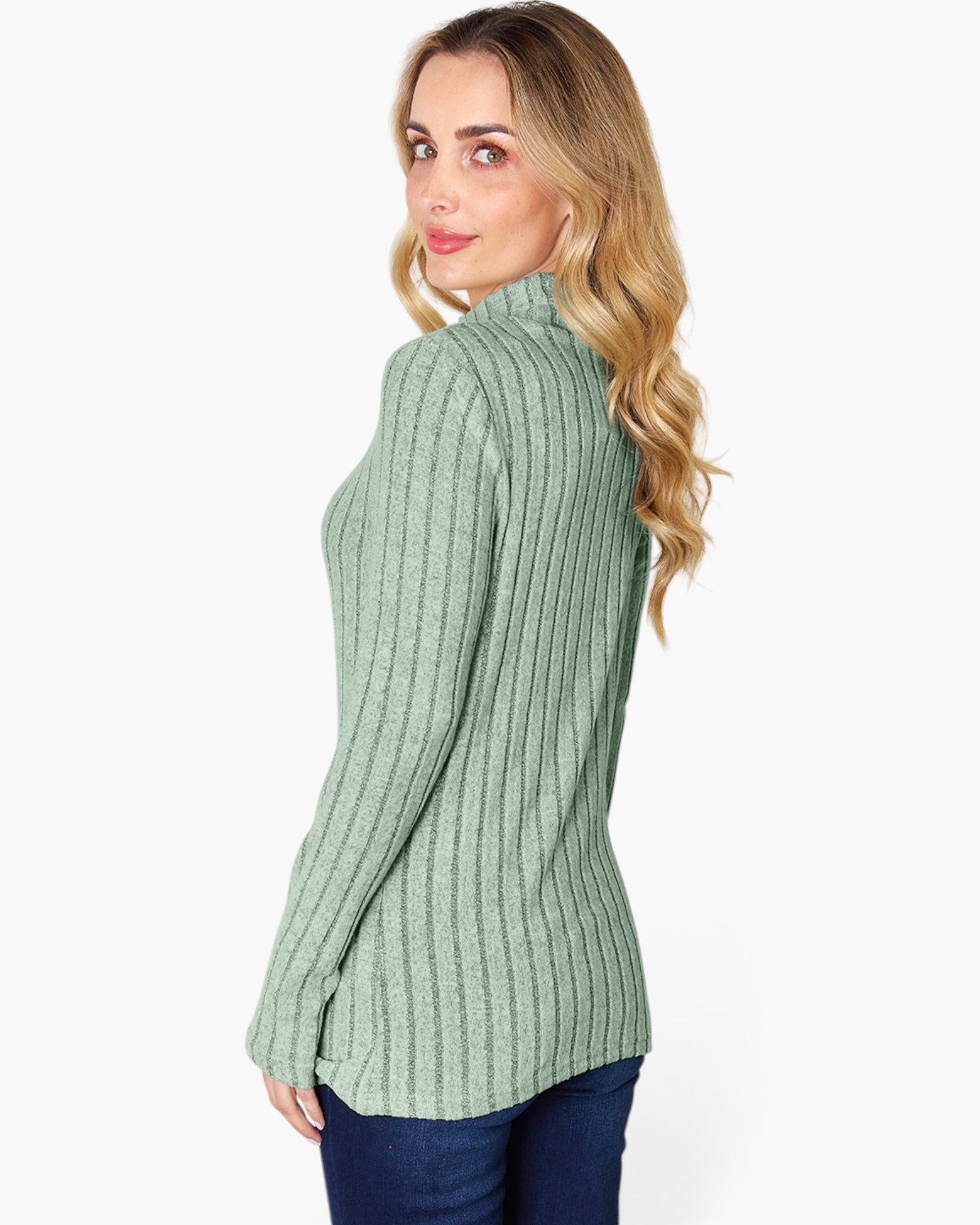 A woman with blonde wavy hair is shown from a side-back angle wearing a sage green ribbed mock neck sweater and dark blue jeans against a plain white background.