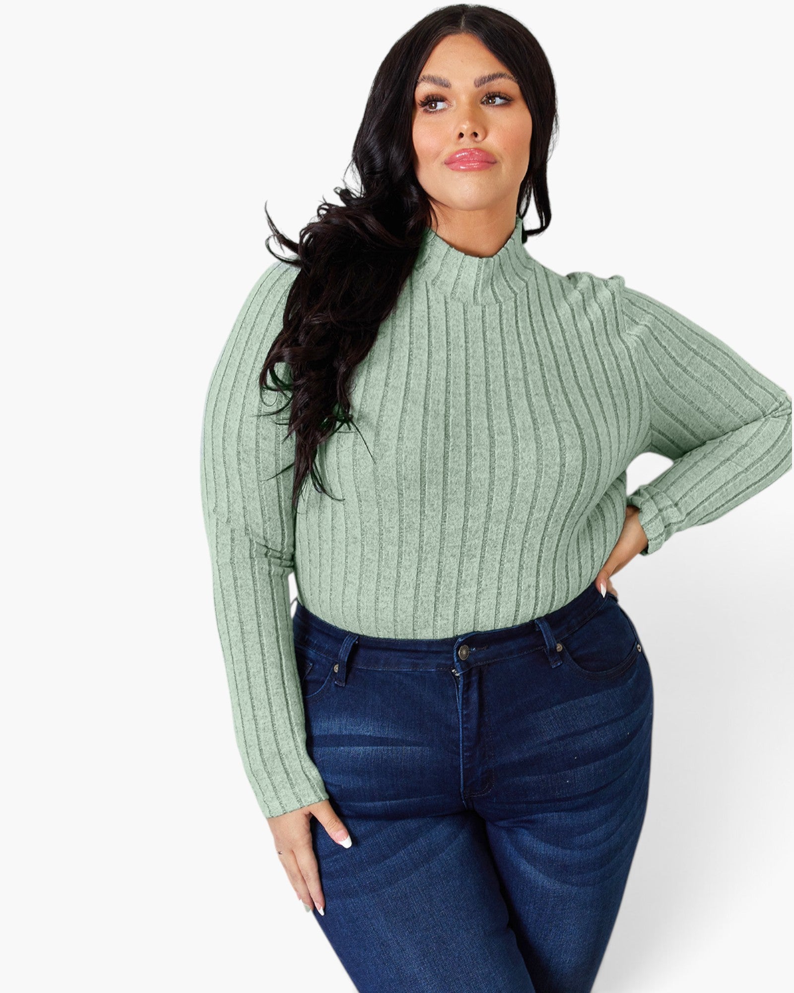 A woman modeling a sage green ribbed mock neck sweater paired with dark blue jeans, photographed against a clean white studio background.