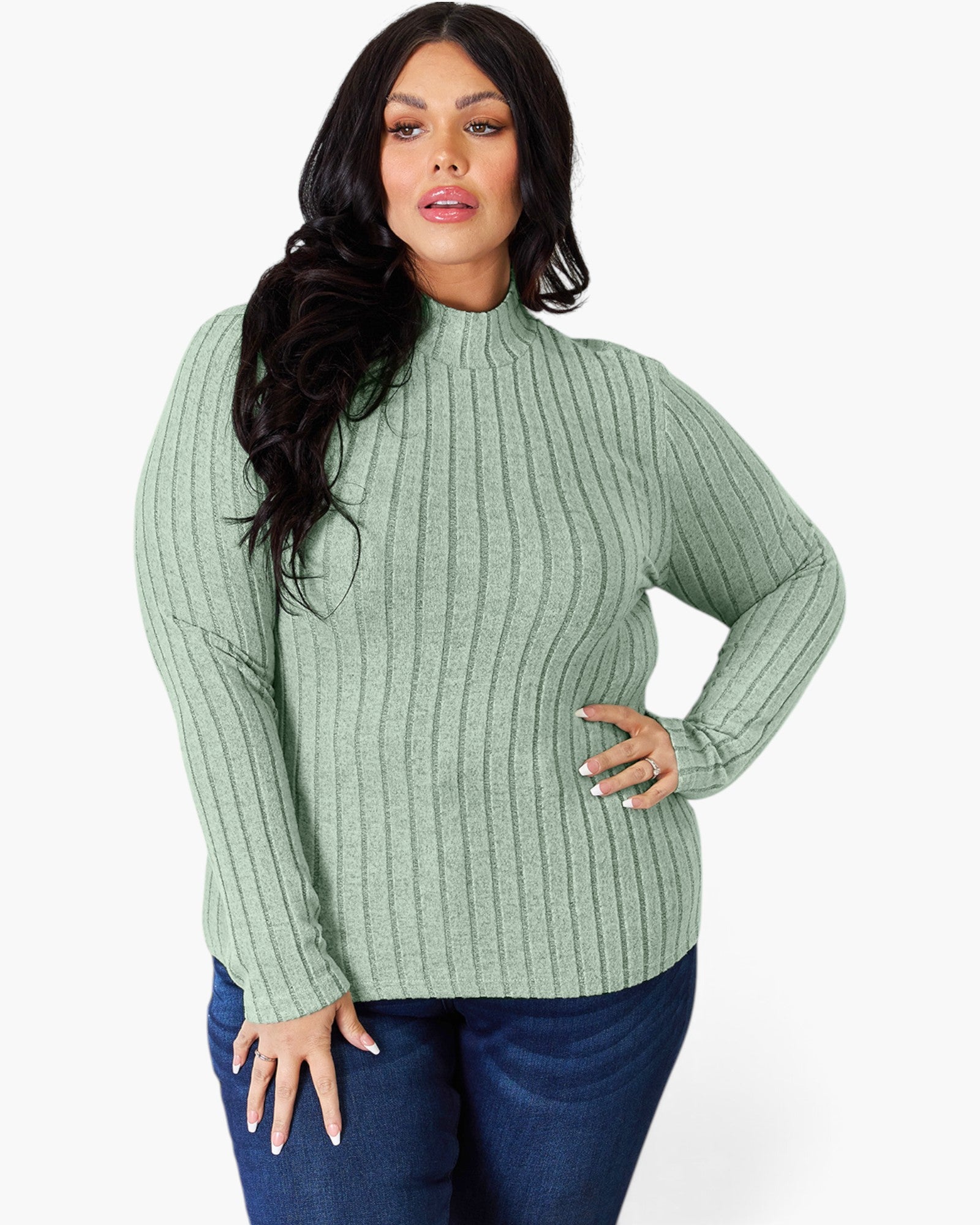 A woman modeling a sage green ribbed mock neck sweater with long sleeves, paired with dark blue jeans, against a plain light gray background.