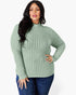 A woman modeling a sage green ribbed mock neck sweater with long sleeves, paired with dark blue jeans, against a plain light gray background.