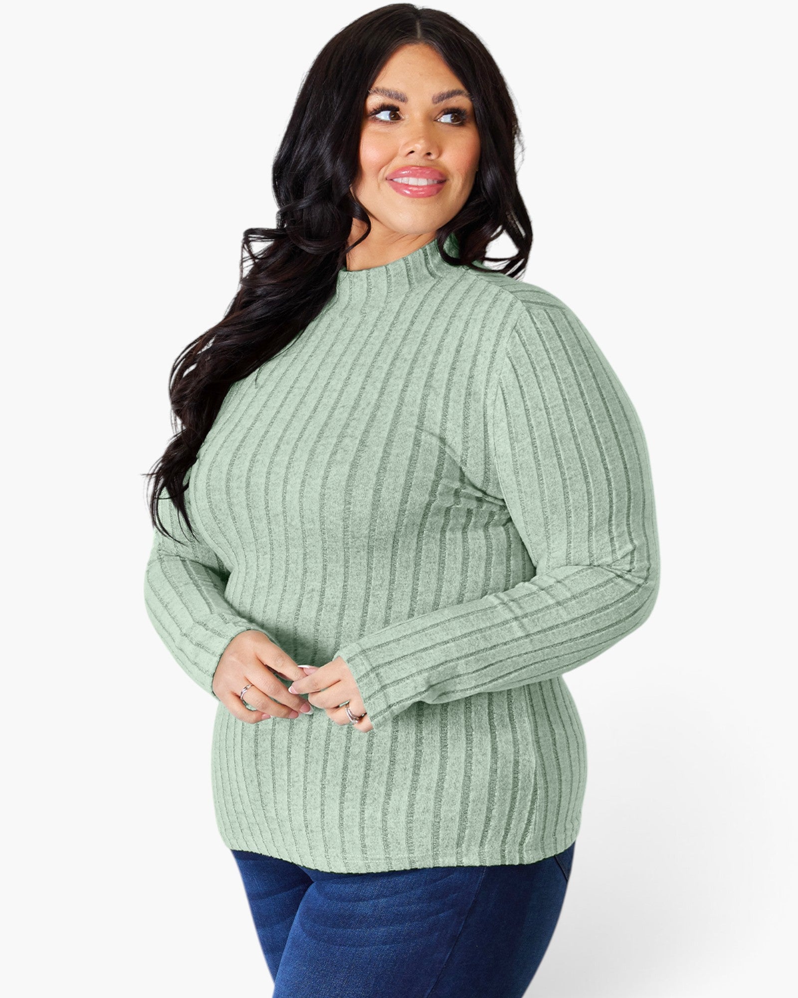 A woman modeling a ribbed knit mock neck sweater in sage green, paired with dark blue jeans, against a plain white studio background.