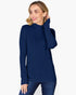 A woman wearing a navy blue ribbed mock neck sweater and dark blue jeans, posing against a light gray studio background.