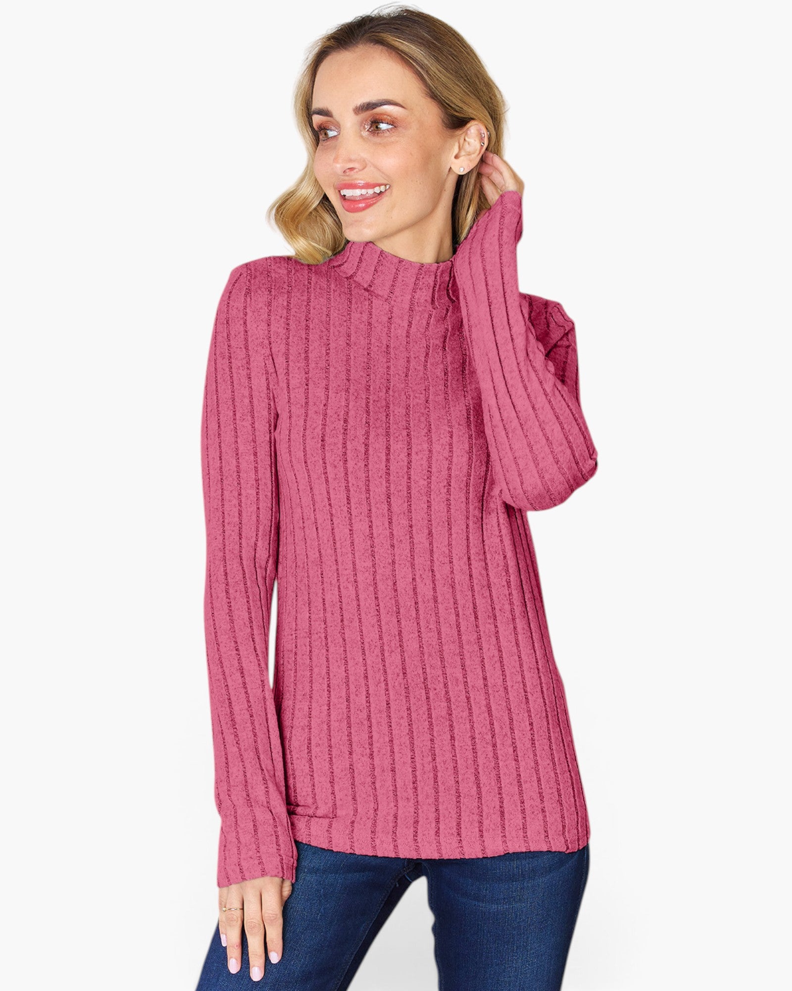 A woman wearing a pink ribbed mock neck sweater and blue jeans, posing against a light gray studio background.