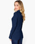 A woman with blonde wavy hair is shown from the side/back wearing a navy blue ribbed knit mock neck sweater and dark blue jeans against a plain white studio background.