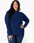 A woman modeling a navy blue ribbed knit mock neck sweater with long sleeves, paired with dark blue jeans, against a plain light gray background.