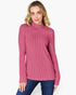 A woman wearing a ribbed pink mock neck sweater and blue jeans, standing against a light gray studio background.