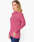 A woman wearing a pink ribbed mock neck long-sleeve top and blue jeans, shown from a side profile against a light gray studio background.