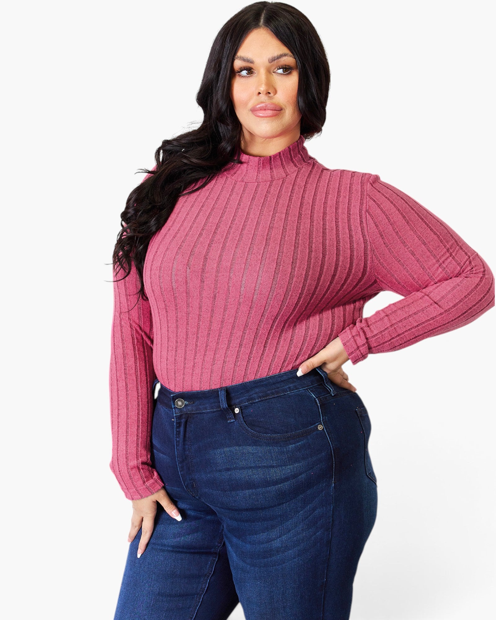 A woman modeling a long-sleeve ribbed mock neck top in dusty rose pink, paired with dark blue jeans, against a plain white studio background.