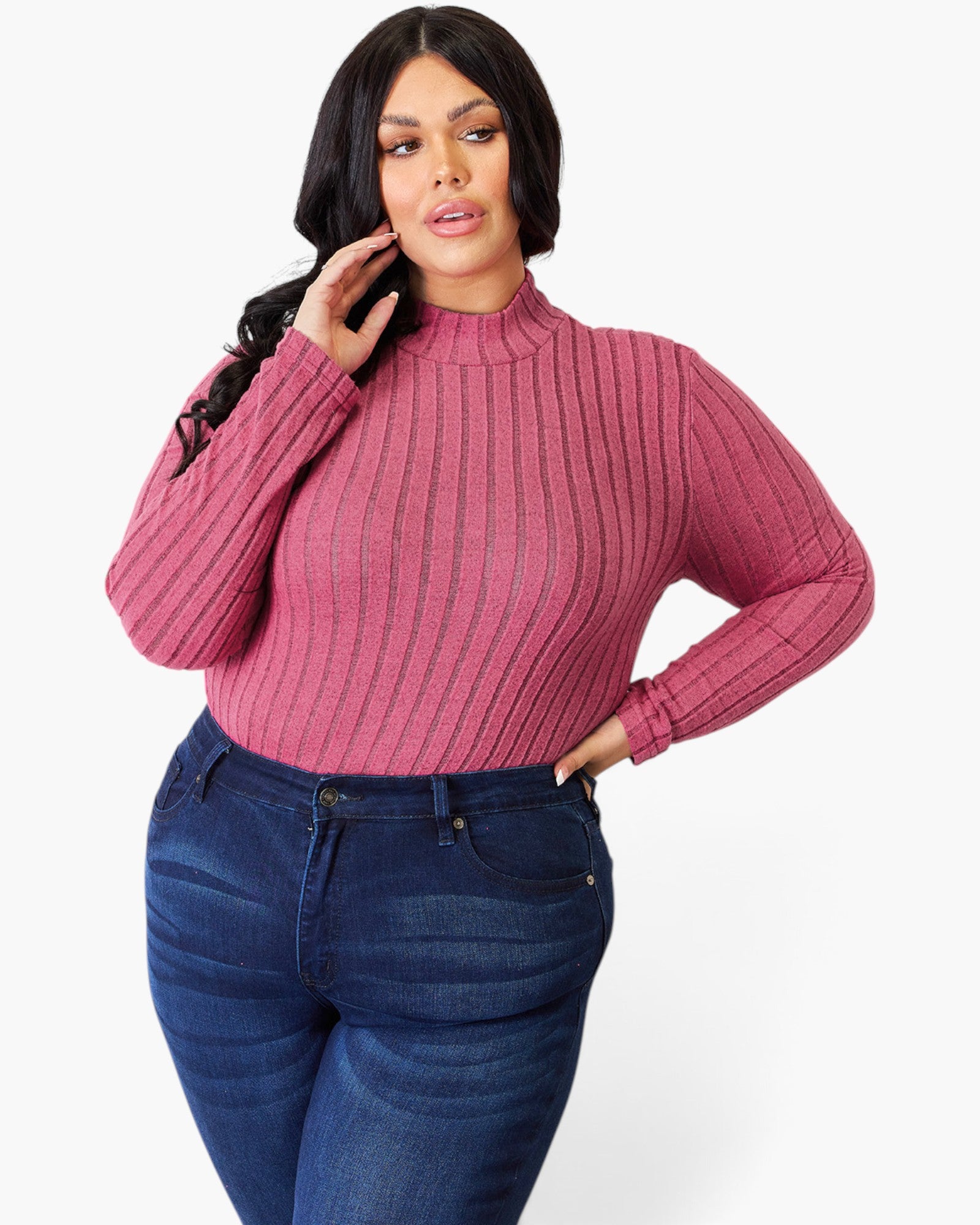 A woman modeling a ribbed long-sleeve mock neck sweater in dusty rose pink, paired with dark blue jeans, against a plain white studio background.