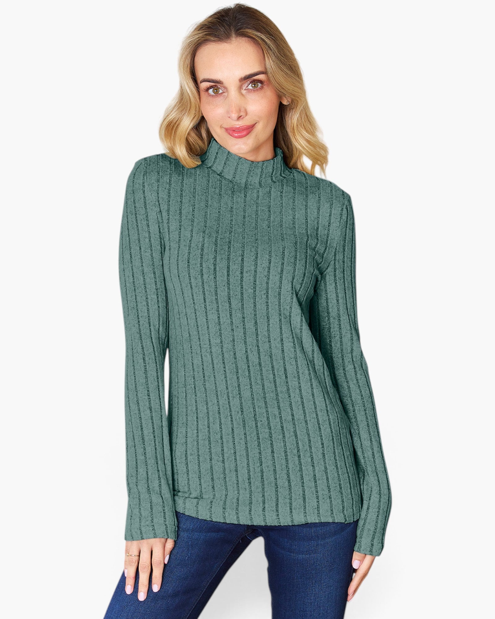 A woman wearing a ribbed knit mock neck sweater in a dusty sage green color, paired with dark blue jeans, against a plain white background.