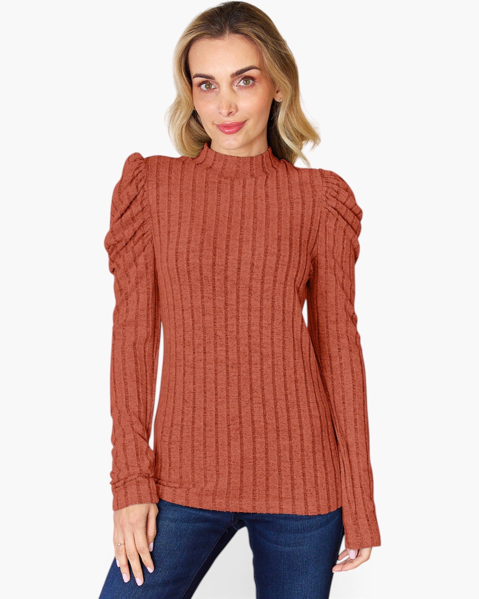 A woman wearing a rust-orange ribbed knit mock-neck sweater with puffed sleeves, paired with blue jeans, against a plain white background.