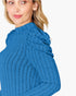 A close-up of a woman wearing a bright blue ribbed knit sweater with a high mock neck and textured shoulder detailing, set against a plain white background.