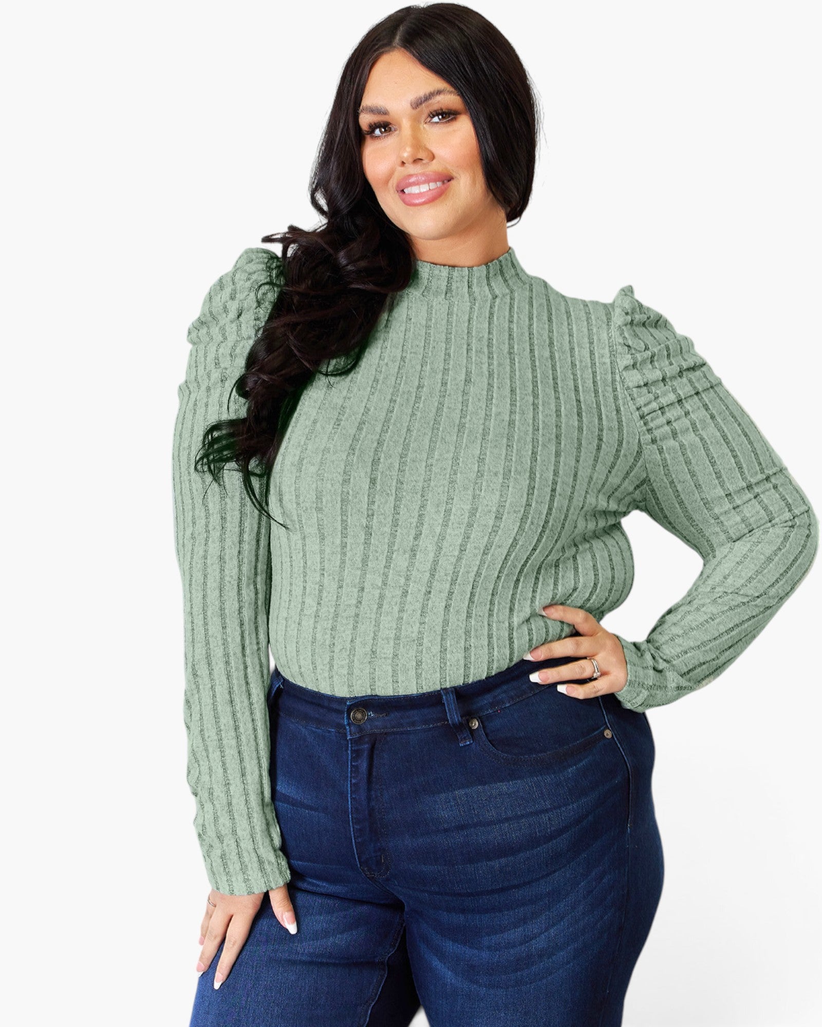 A woman modeling a sage green ribbed knit mock neck sweater with puffed shoulders, paired with dark blue jeans, against a plain white background.