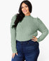 A woman modeling a sage green ribbed knit mock neck sweater with puffed shoulders, paired with dark blue jeans, against a plain white background.