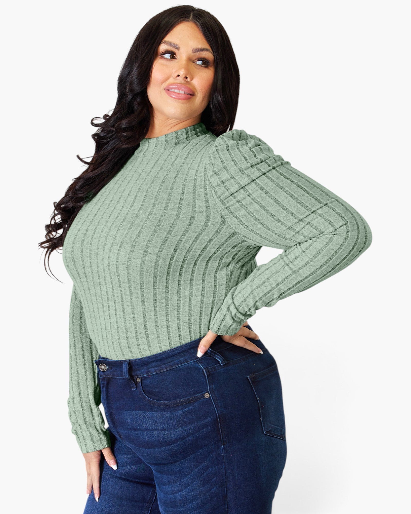 A woman modeling a sage green ribbed knit mock neck sweater with balloon sleeves, paired with dark blue jeans, against a plain white background.