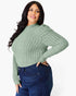 A woman modeling a sage green ribbed knit mock neck sweater with balloon sleeves, paired with dark blue jeans, against a plain white background.