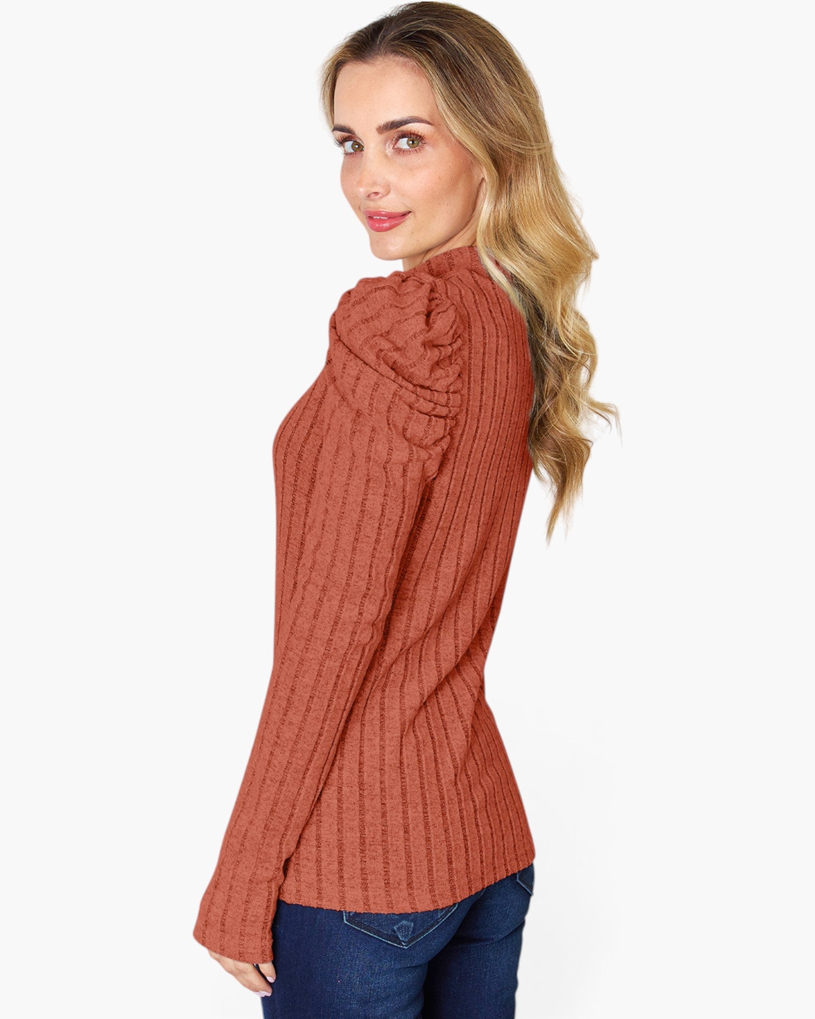 A woman wearing a ribbed knit long-sleeve sweater in a warm rust or terracotta color, paired with blue jeans, shown from a side-back angle against a plain light gray studio background.