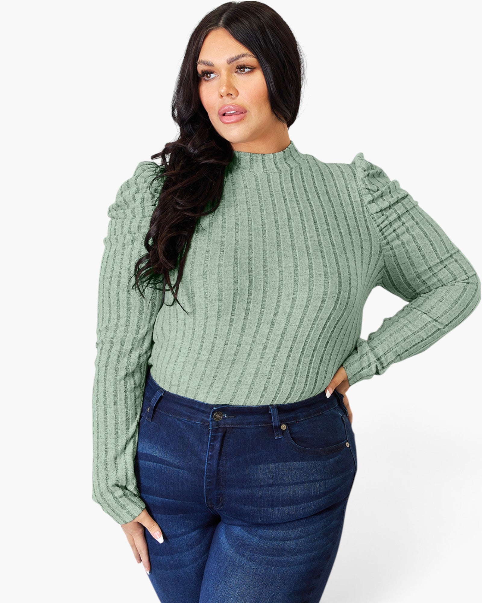 A woman modeling a sage green ribbed knit mock neck top with ruffled long sleeves, paired with dark blue jeans, against a plain white studio background.