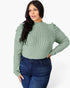 A woman modeling a sage green ribbed knit mock neck top with ruffled long sleeves, paired with dark blue jeans, against a plain white studio background.