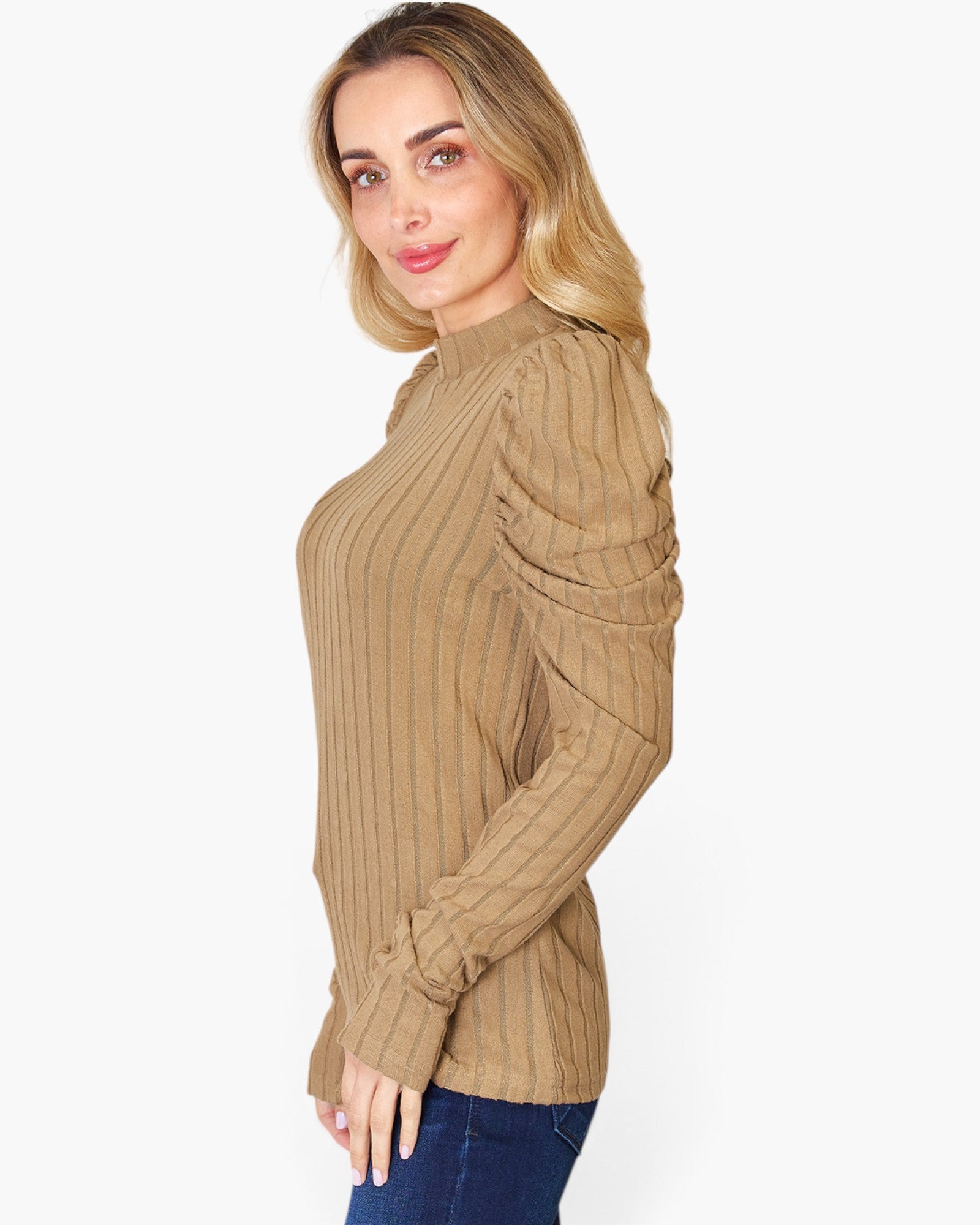 A woman modeling a tan ribbed mock neck sweater with ruched sleeves, paired with blue jeans, against a plain white background.