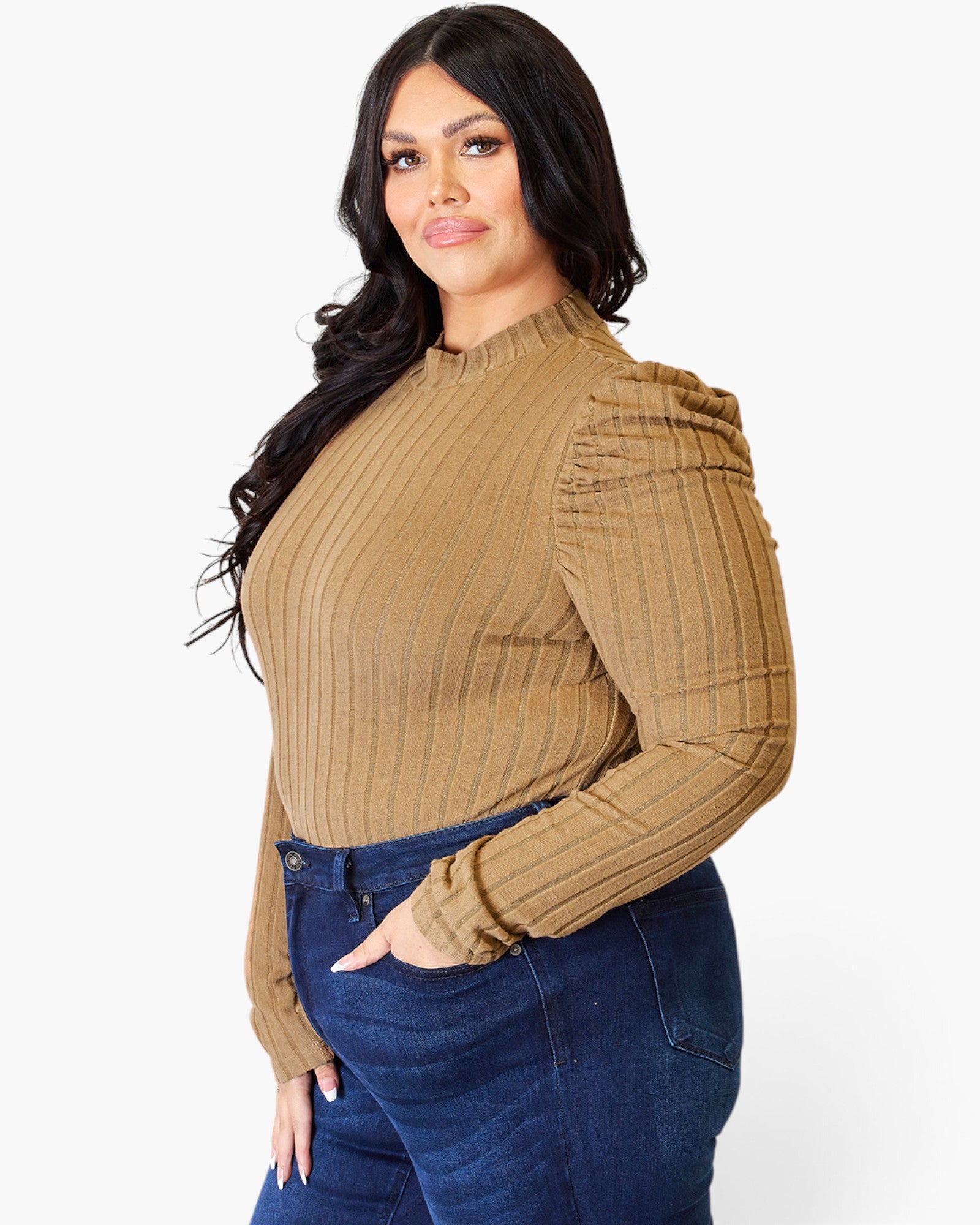A woman wearing a tan ribbed long-sleeve mock neck top with puffed shoulders and dark blue jeans, photographed against a light gray studio background.
