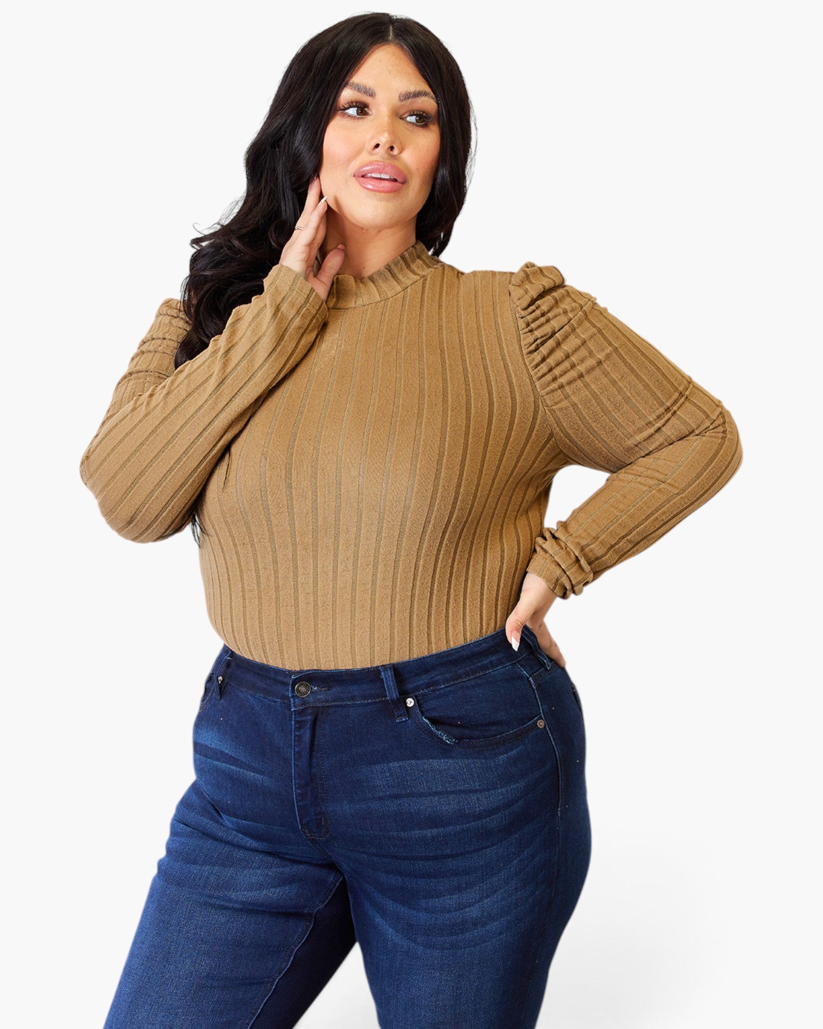 A woman modeling a ribbed knit long-sleeve top in camel tan with puffed shoulders, paired with dark blue jeans, against a plain light gray studio background.