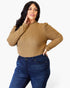 A woman modeling a ribbed knit long-sleeve top in camel tan with puffed shoulders, paired with dark blue jeans, against a plain light gray studio background.