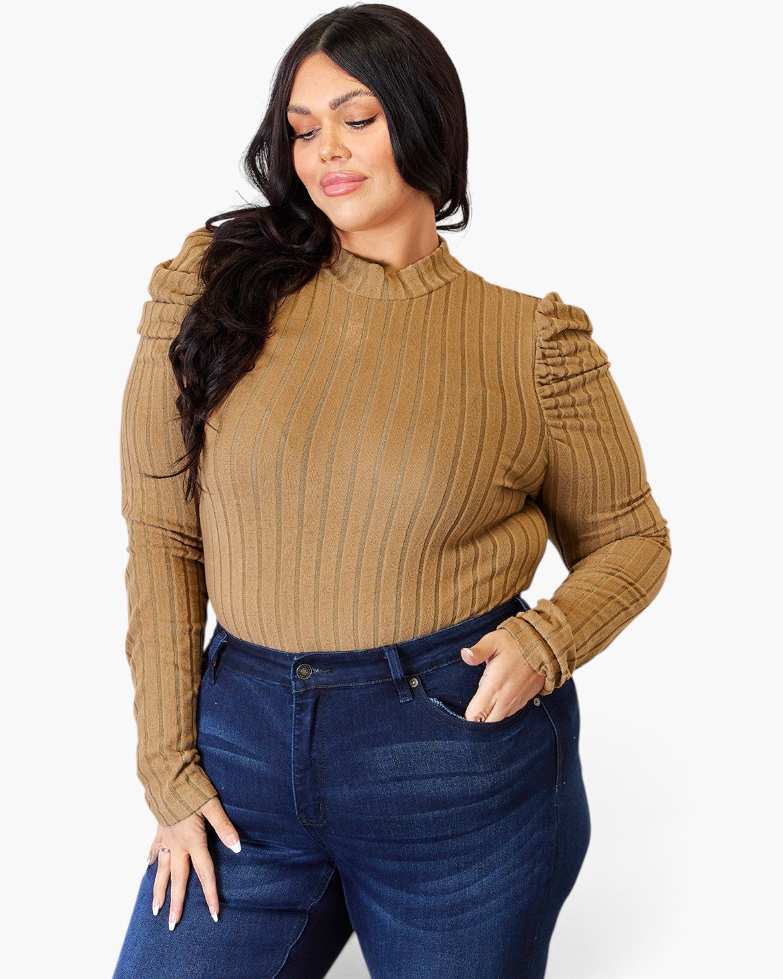 A woman wearing a tan ribbed mock neck long-sleeve top with ruched shoulders and dark blue jeans, standing against a light gray studio background.