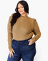 A woman wearing a tan ribbed mock neck long-sleeve top with ruched shoulders and dark blue jeans, standing against a light gray studio background.