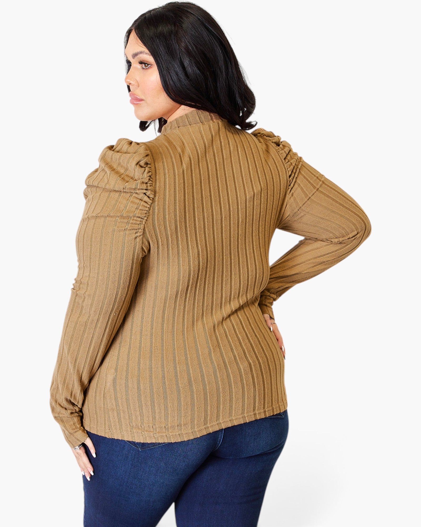 A woman shown from the back wearing a tan ribbed long-sleeve blouse with ruched shoulder details and dark blue jeans, against a plain light gray studio background.