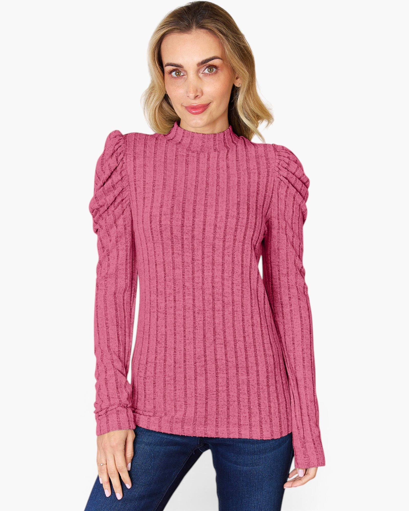 A woman wearing a pink ribbed knit mock-neck sweater with dramatic shoulder detailing, paired with blue jeans, against a plain light gray background.