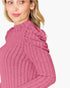 A close-up of a woman wearing a ribbed knit mock neck sweater with dramatic ruffled detailing on the shoulders in a dusty rose pink color, set against a plain white background.