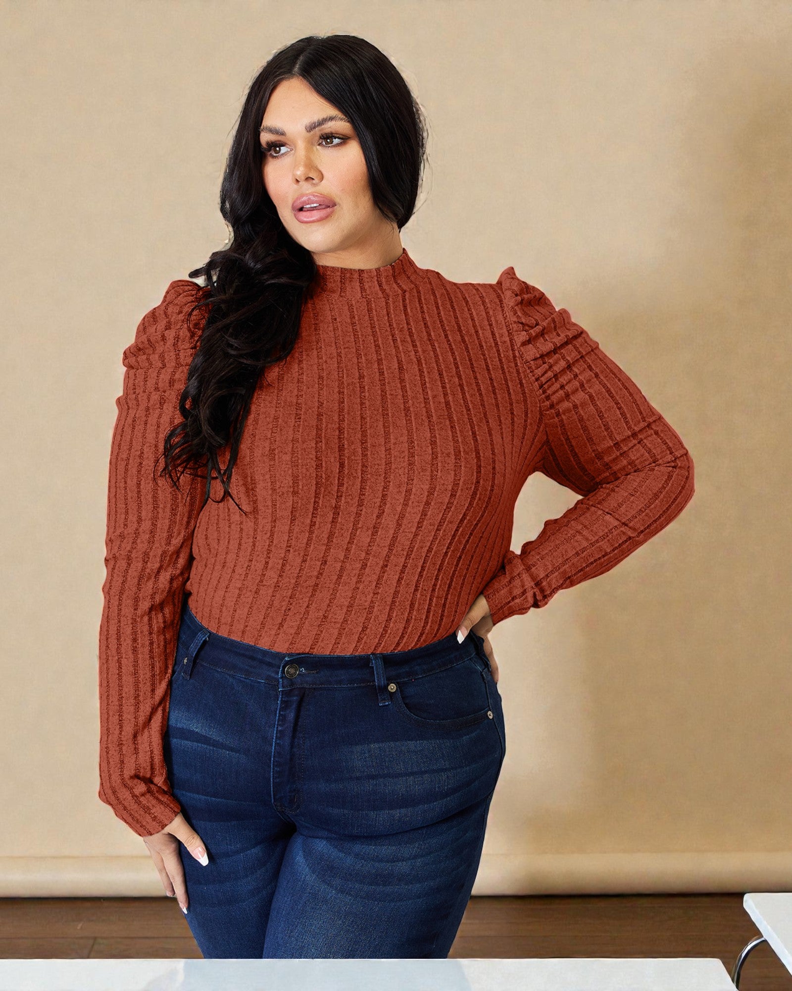 Woman wearing a rust-colored ribbed sweater and blue jeans against a beige background