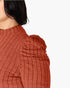Close-up of a person wearing a ribbed knit top in a warm rust or terracotta color, showing the shoulder and side of the head against a plain light background.