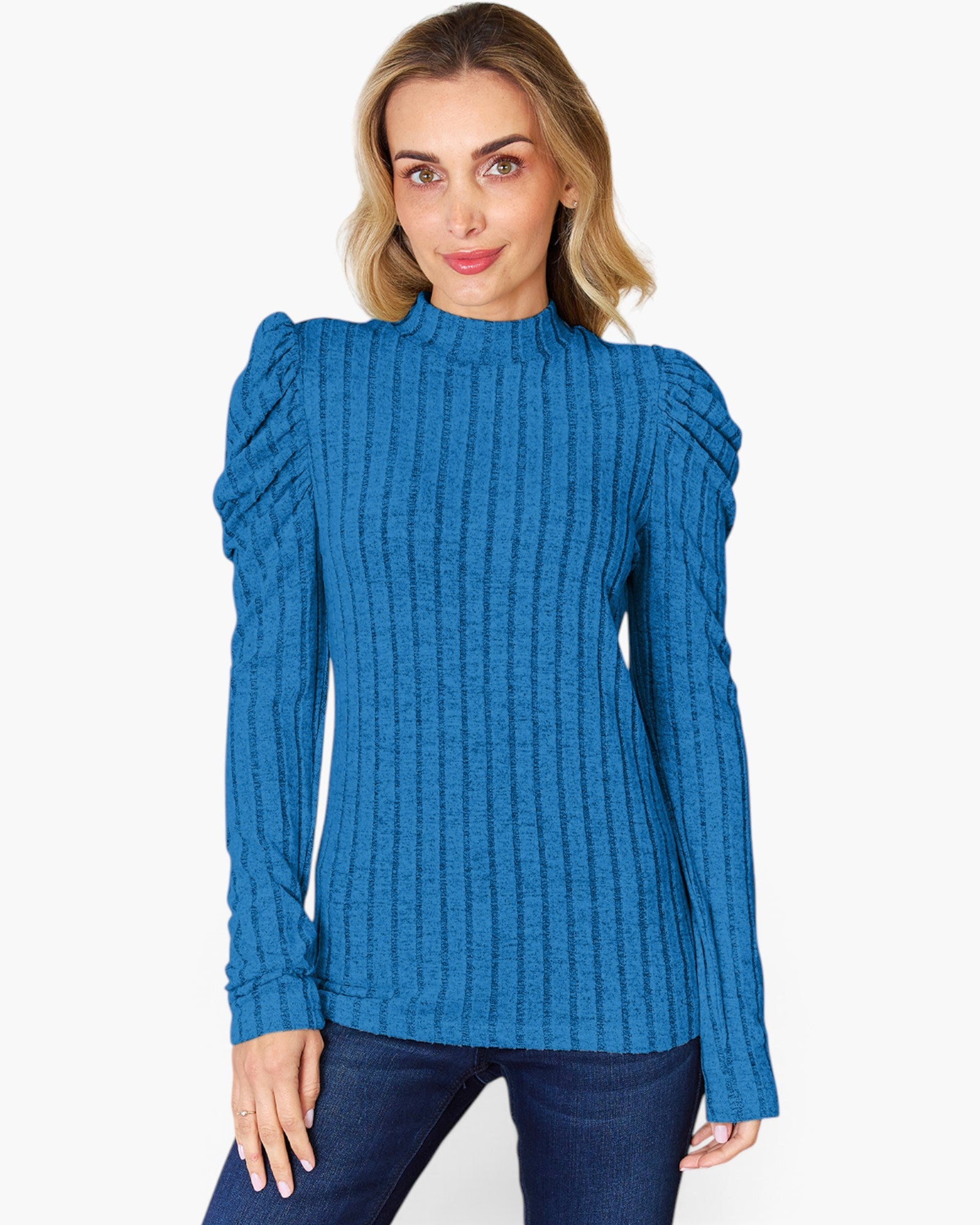 A woman wearing a blue ribbed knit mock-neck sweater with dramatic puffed shoulders, paired with dark blue jeans, against a plain light gray background.