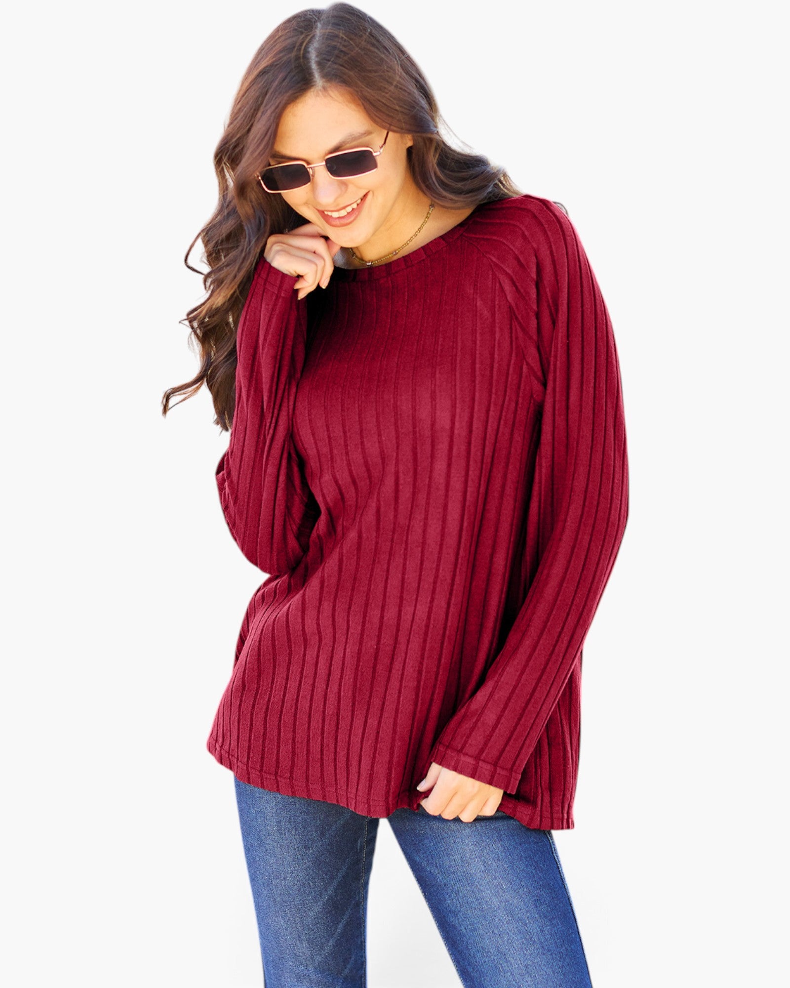 A woman wearing a ribbed knit long-sleeve sweater in deep burgundy, paired with blue jeans and sunglasses, against a plain white background.