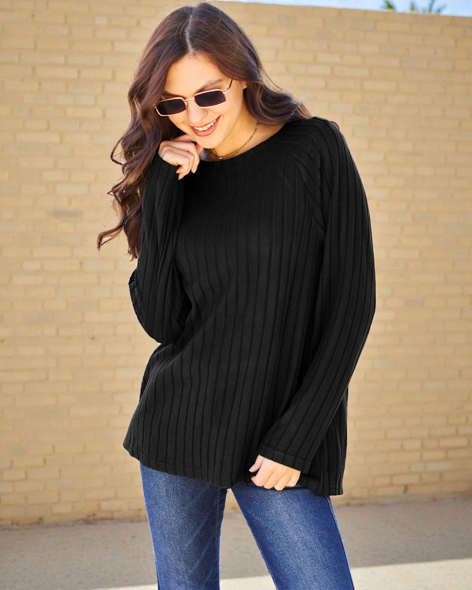 Woman wearing a black sweater and blue jeans against a beige brick wall.